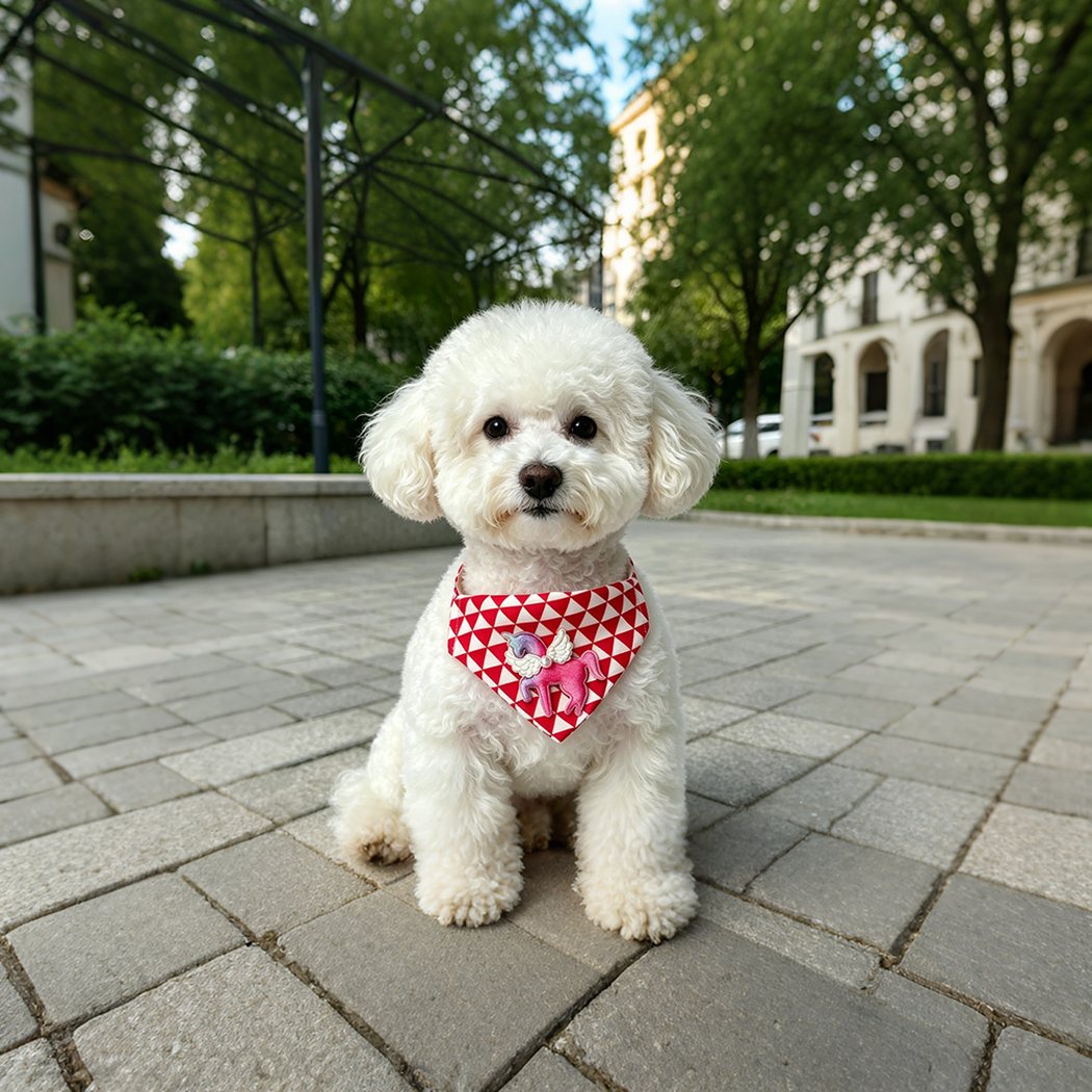 White dog sitting on a paved walkway wearing red-and-white Bandana pet neckwear with a winged unicorn accent.