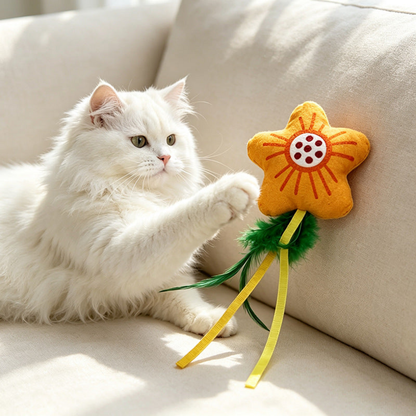 Flower cat toy with green feather-like tails and yellow ribbons beside a white cat on a sofa