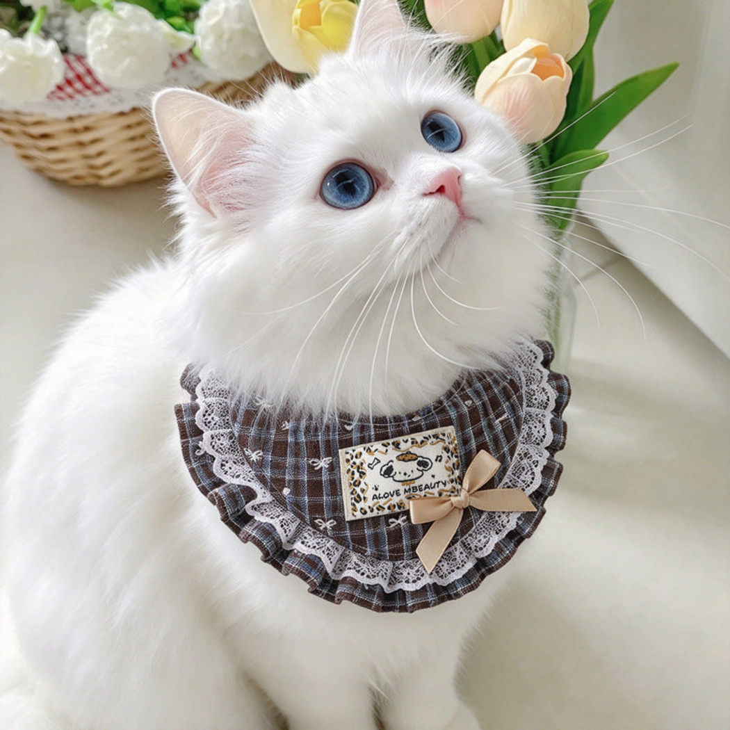 Brown plaid Pet Neckwear with white lace-look edging and a beige bow, worn by a white cat beside flowers