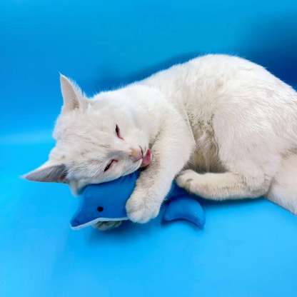 White cat lying beside the Dolphin cat toy with its tongue near the toy and a front leg draped over it