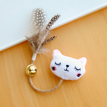 White cat toy with embroidered closed eyes, pink cheeks, a braided cord, gold-colored bell and spotted feathers.
