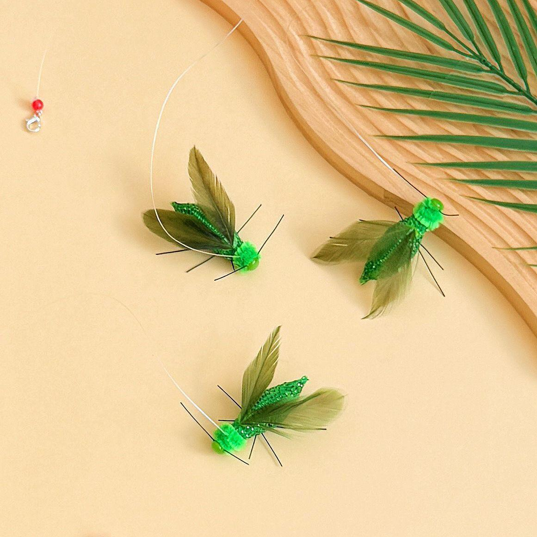 Three green winged insect-shaped cat toy attachments arranged with a leaf and wood-look prop