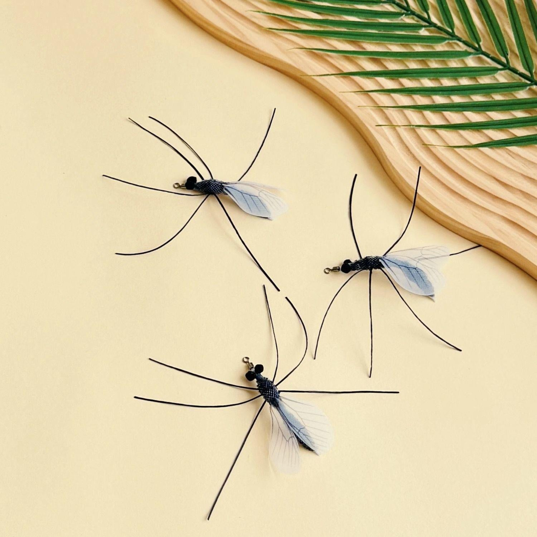 Three mosquito-shaped cat toy attachments arranged beside a woven tray and palm leaf