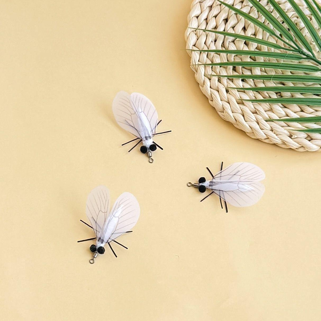Three white moth-shaped cat toys arranged beside a woven mat and green leaves