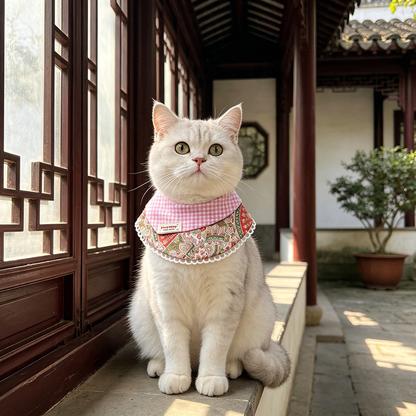 A cat wearing pink gingham and floral pet neckwear while sitting beside a window.