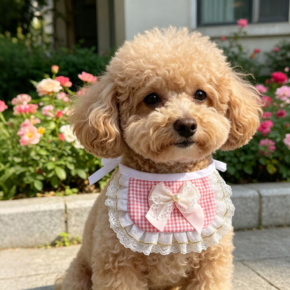 Tan dog wearing pink gingham pet neckwear with a white bow and ruffled edging beside flowering plants.