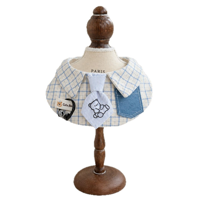 Blue and cream checked pet neckwear on a display stand, showing the curved bib shape, bear tie, and blue pocket.